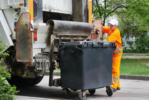 A worker dressed in high-visibility orange workwear and a white safety helmet is standing next to a large black wheeled rubbish bin on a street or driveway, near a waste collection vehicle used for rubbish removal. The worker appears to be overseeing or guiding the bin as it is aligned close to the vehicle’s mechanical lifting arm, which is positioned to load the bin into the vehicle’s hatch. The background features a glimpse of green trees and foliage, indicating an outdoor setting, possibly within a residential or urban area in Clapham. The waste collection truck shows visible metallic components, textured surfaces, and a partially visible conveyor or roller mechanism used for lifting and depositing rubbish. The scene is illuminated by natural daylight, emphasizing the clean, organized environment typical of professional rubbish removal services provided by Clapham House Clearance, which operates across local London areas, including postcode regions near Clapham.