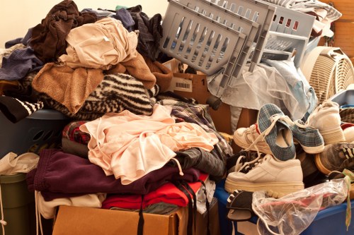 A large collection of assorted clothing, fabric, and textiles piled on top of each other, including shirts, trousers, and other garments in various colours and materials. The pile also contains some footwear such as sneakers and sandals, along with plastic bags, cardboard boxes, and laundry baskets scattered throughout. The scene appears to be set indoors, possibly in an unorganized storage or staging area for rubbish removal, with the items occupying a significant portion of the space. This dense accumulation of clothing and household items is typical of waste cleared during residential rubbish removal services in areas such as Clapham, suggesting a need for professional clearance by a company like Clapham House Clearance.