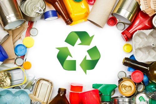 A diverse assortment of discarded items arranged in a circular pattern on a white background, featuring metal cans, plastic bottles, cardboard tubes, and paper packaging with various colors including silver, blue, red, yellow, and brown. At the centre of the arrangement is a large, green recycling symbol consisting of three arrows forming a triangle, symbolising waste reuse and recycling efforts. The objects include beverage cans with ridged surfaces, plastic bottles with caps, and cardboard tube sections, all appearing used and crumpled, illustrating common household rubbish. The lighting is bright and even, emphasizing the textures and colours of the waste materials. This image visually supports rubbish removal and waste recycling services offered by Clapham House Clearance, subtly referencing environmental responsibility within a London suburban context, indicated by the postcode inference from the company’s service area. The setting appears to be an indoor or outdoor space suitable for waste collection, emphasizing the importance of sorting and recycling waste efficiently in local communities.