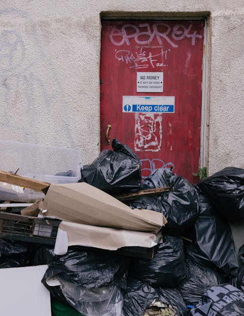 A collection of black garbage bags and miscellaneous waste items piled against an exterior wall in front of a red metal door covered with graffiti and stickers, including a white sign indicating 'No M